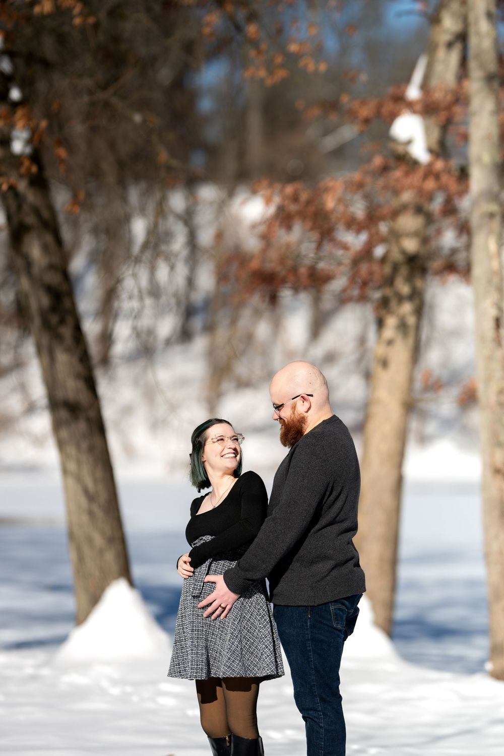 Smiling pregnant woman and bearded partner holding her belly together in a sunny snowy winter park.
