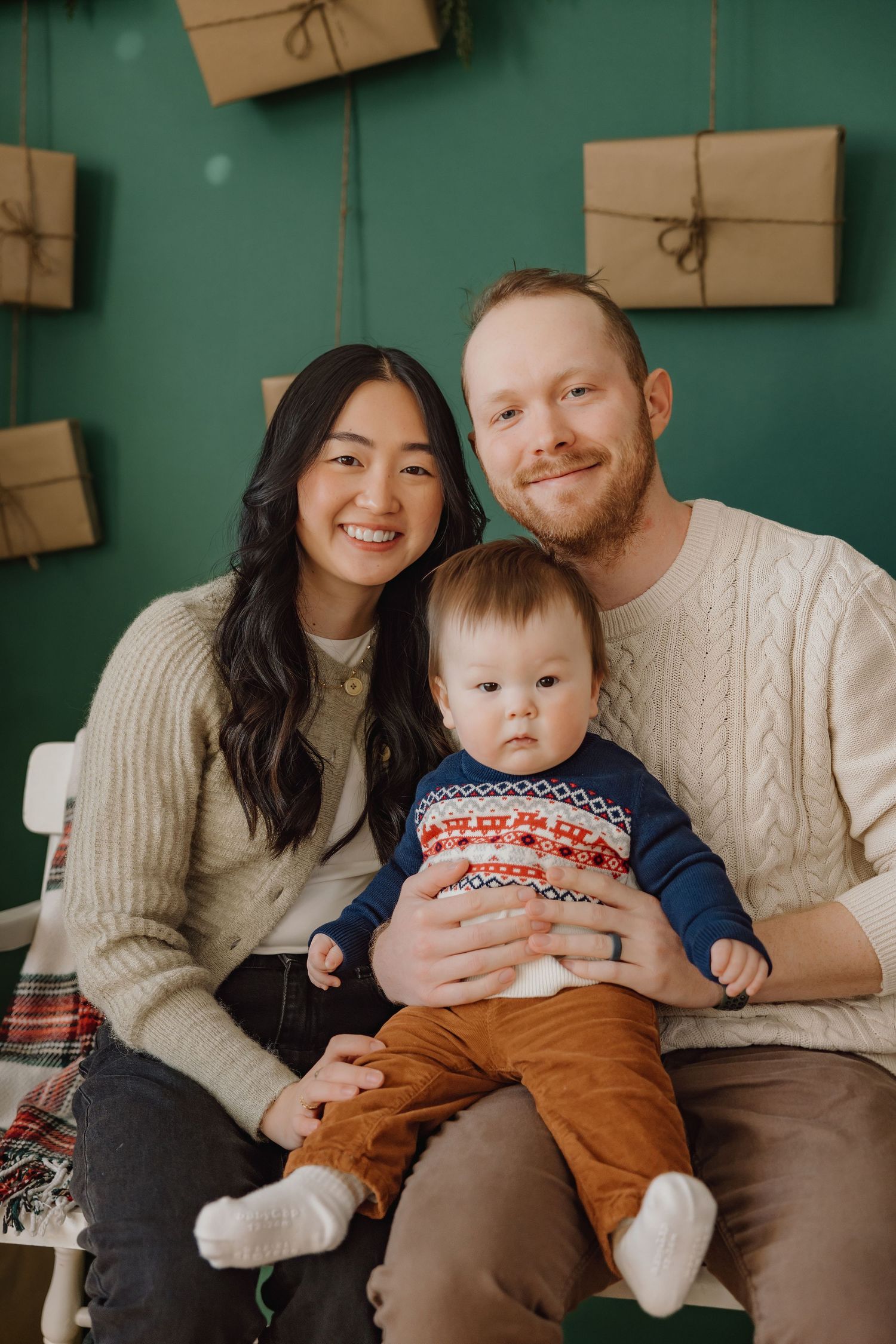 Happy interracial family with redhead father, Asian mother, and baby boy posing before green Christmas backdrop.