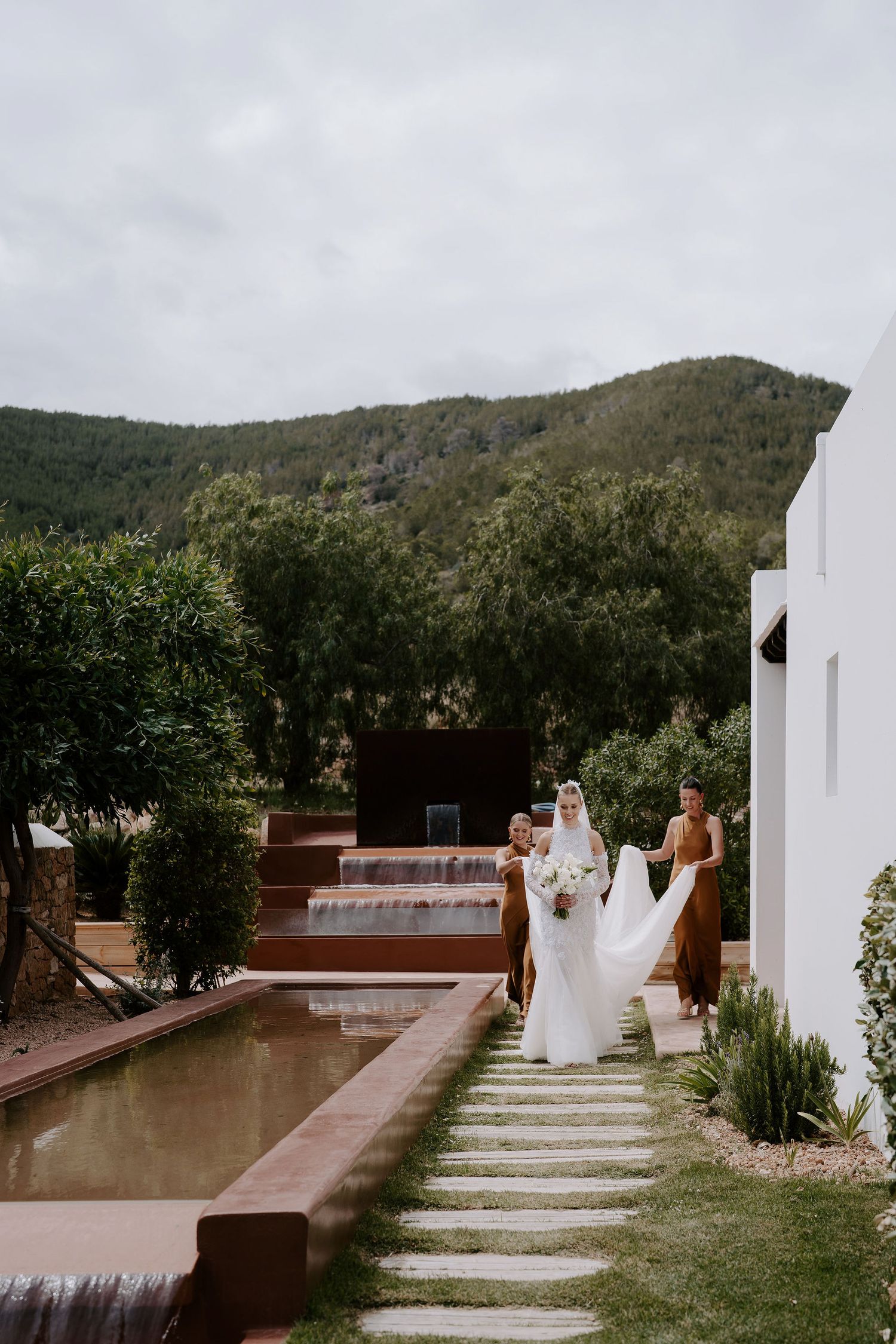 bridesmaids carrying brides Emma Beaumont dress as they walk alongside pond at casa maca ibiza