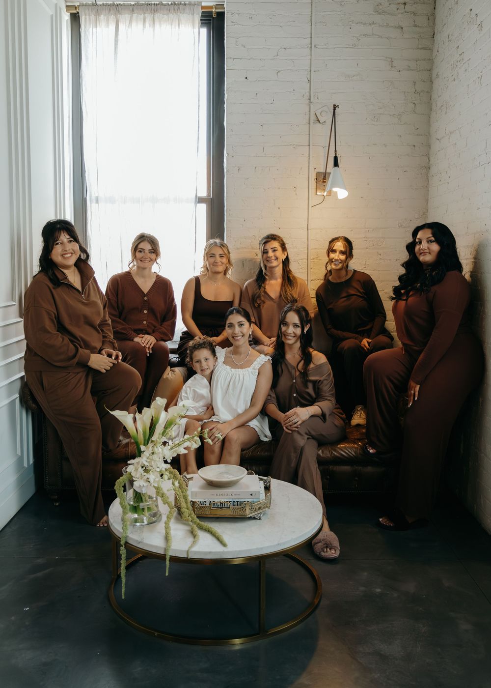 Bridesmaids in brown dresses gather around a modern coffee table in an industrial-style room with white brick walls.