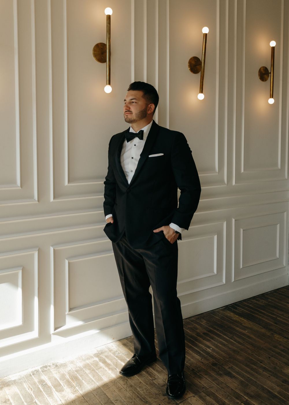 Groomsmen in black tuxedos stand in an elegant room with white paneled walls and hanging pendant lights.