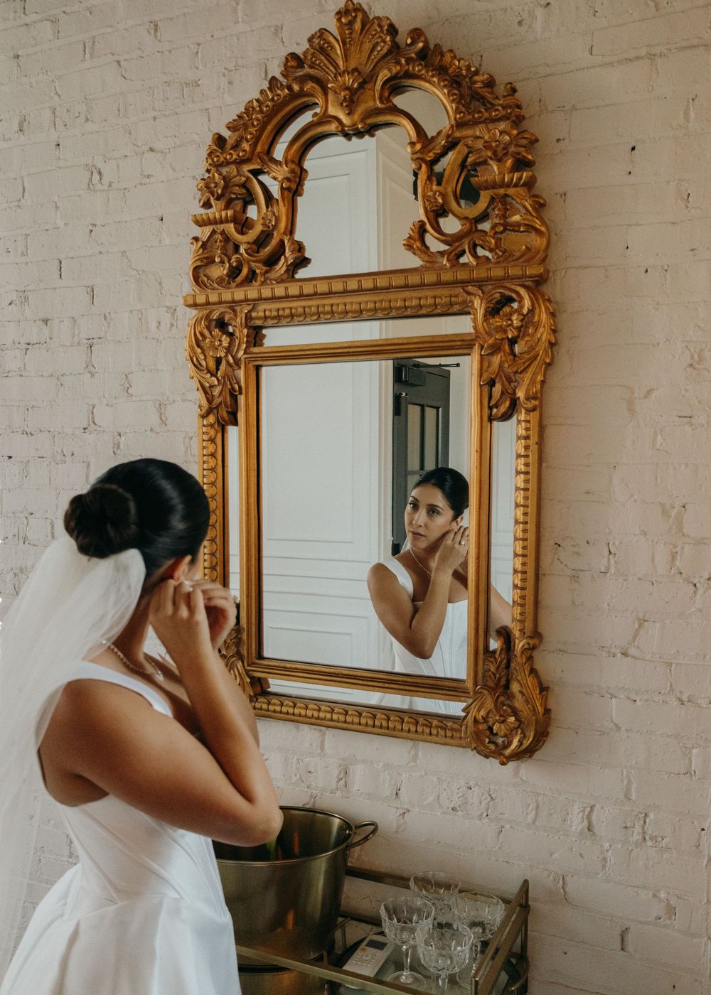 Ornate gold-framed mirrors hang on white brick walls in an elegant getting-ready space.
