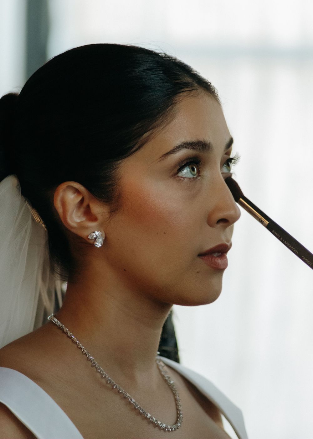 A bride in a white veil and diamond necklace has makeup applied with a brush during wedding preparations.