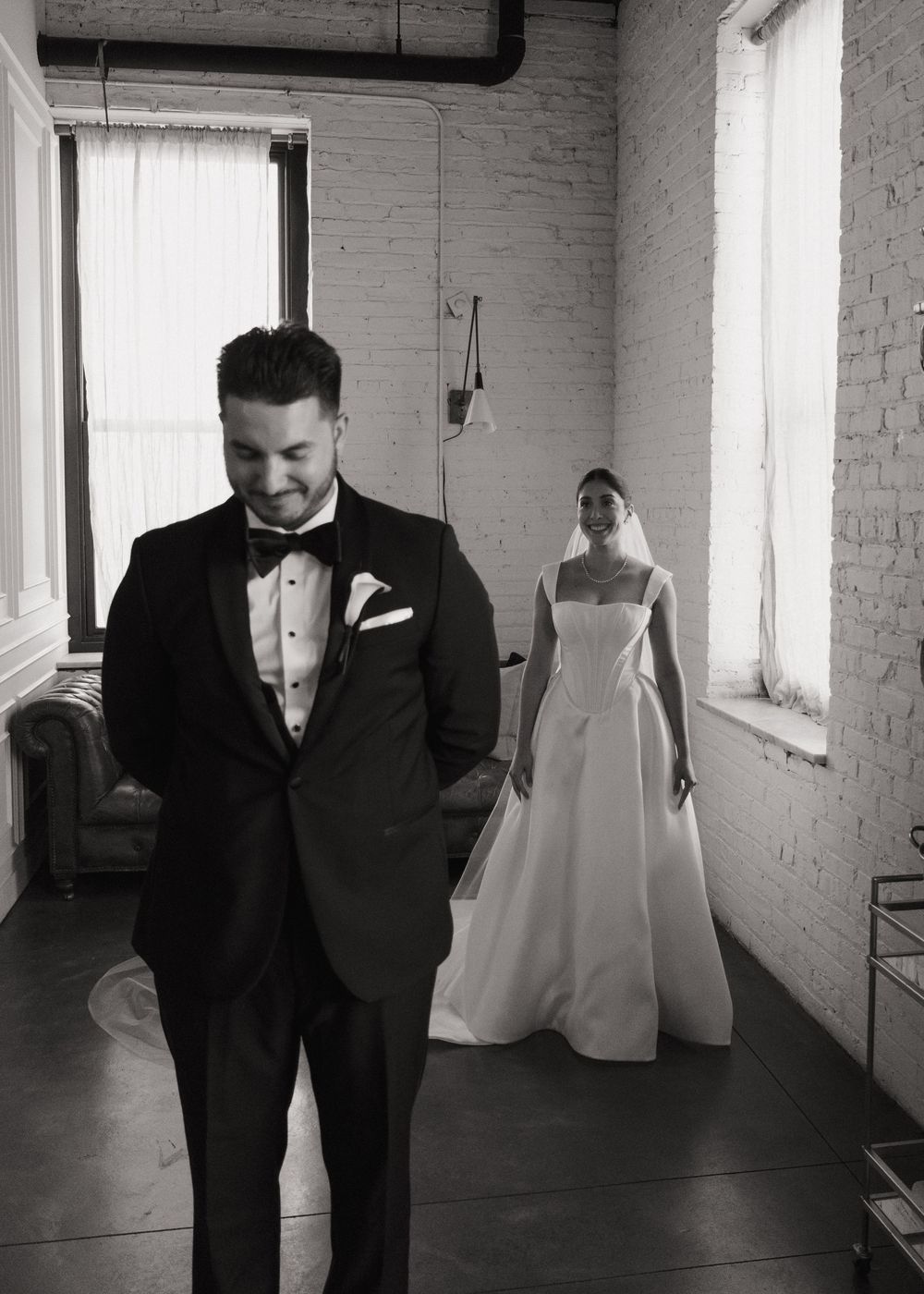 Wedding attire including black tuxedos and white dresses displayed in an industrial loft space with exposed brick walls.