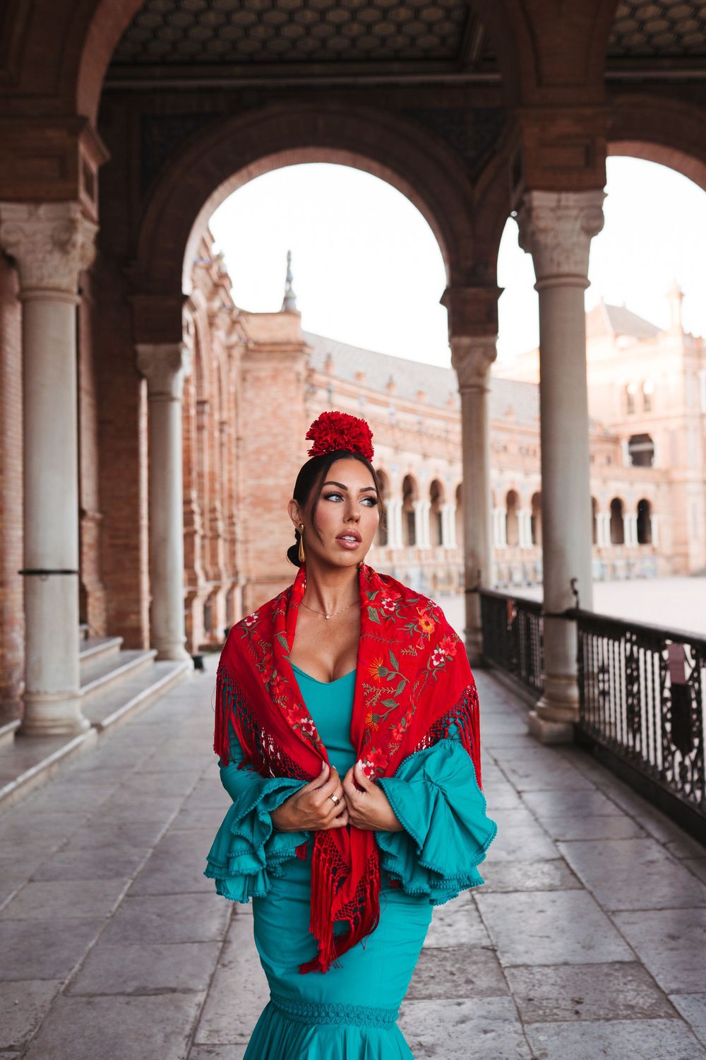 A flamenco dancer in a turquoise dress and red shawl poses under arches at Plaza de España, Seville.