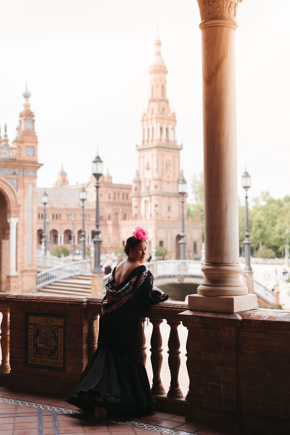 A figure in a black flamenco dress and red flower stands on Plaza de España's ornate balcony in Seville, Spain.