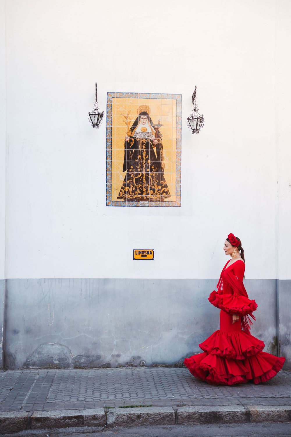 Woman in vibrant red flamenco dress stands against white wall with religious artwork and lanterns in Spain.