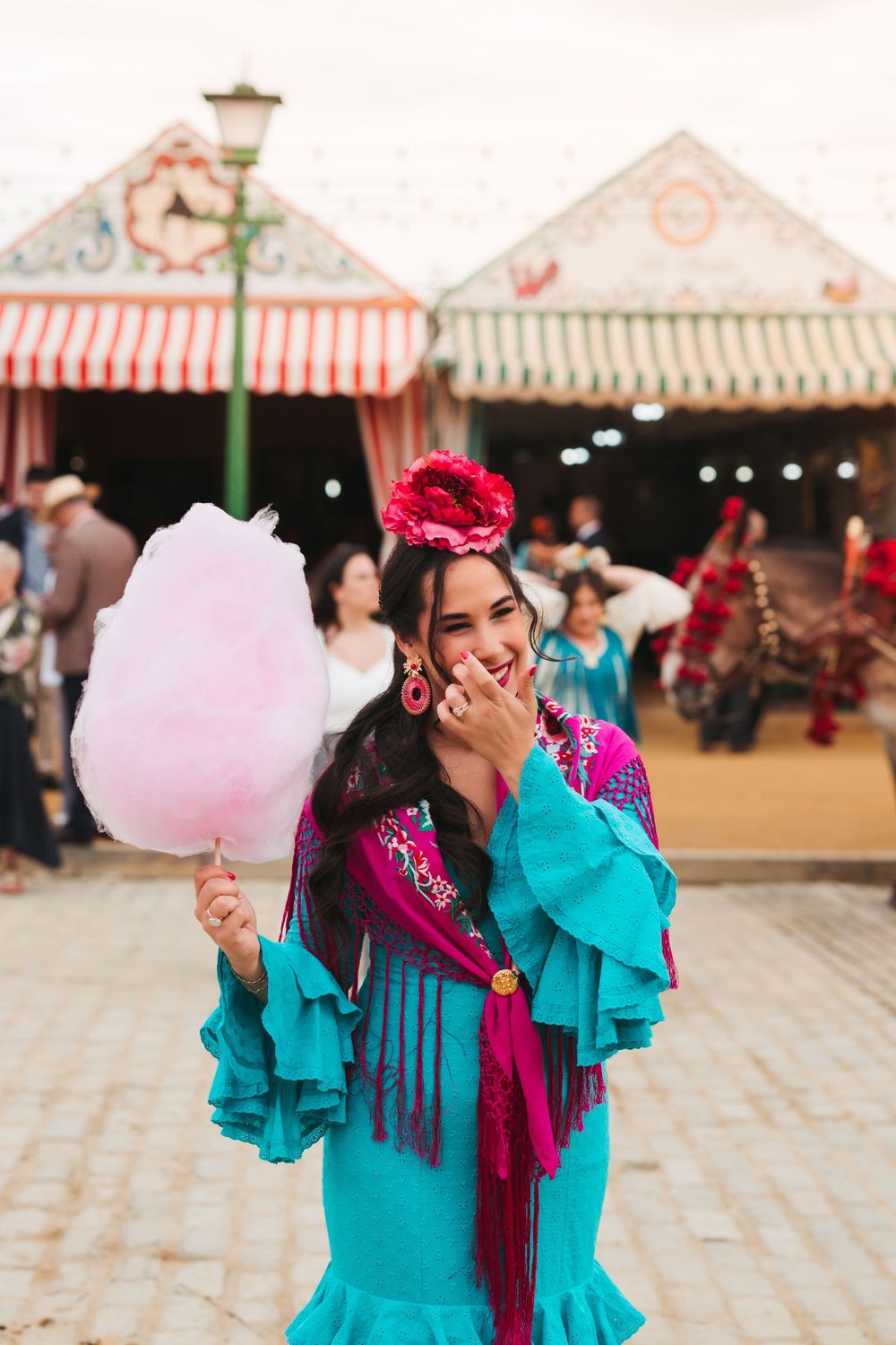 A festive scene at a fair with colorful traditional dress, pink cotton candy, and striped market stalls in the background.