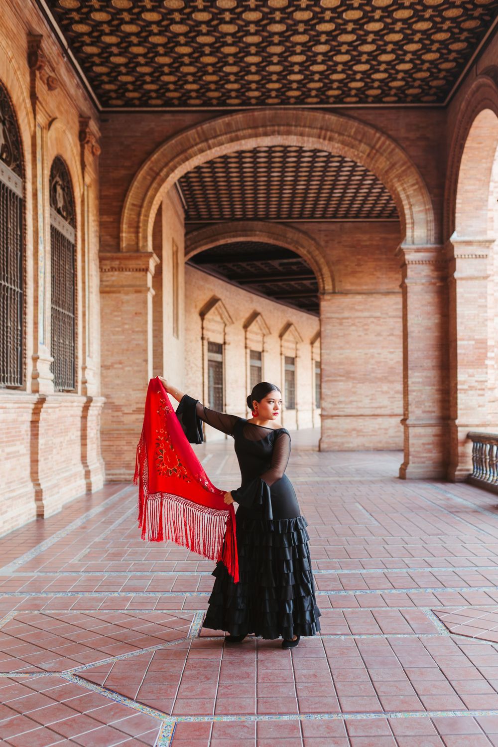 A flamenco dancer performs a dramatic spin with a flowing red shawl in a historic Spanish courtyard with arched columns.
