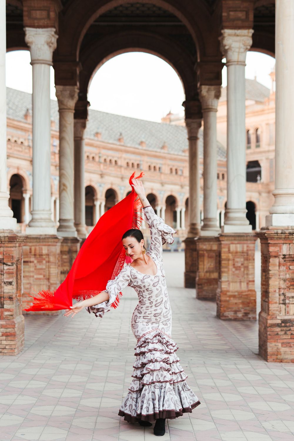 A flamenco dancer in a polka dot dress twirls with a flowing red shawl under ornate archways of Plaza de Espana in Seville.