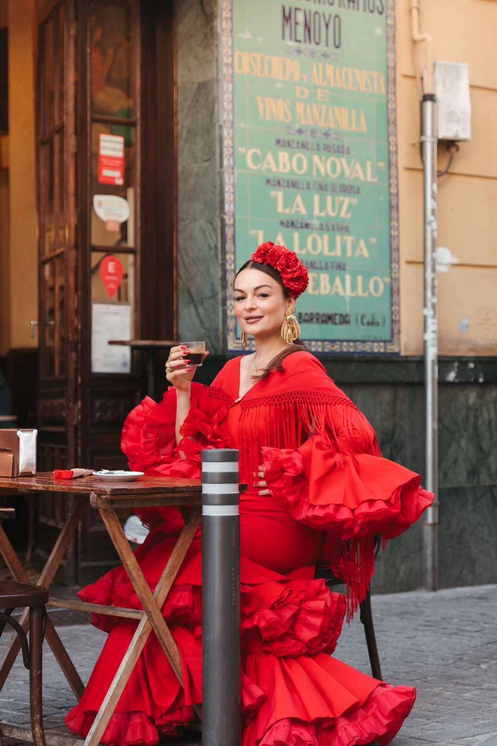 A woman in a vibrant red flamenco dress with ruffles sits at an outdoor cafe table holding a drink.
