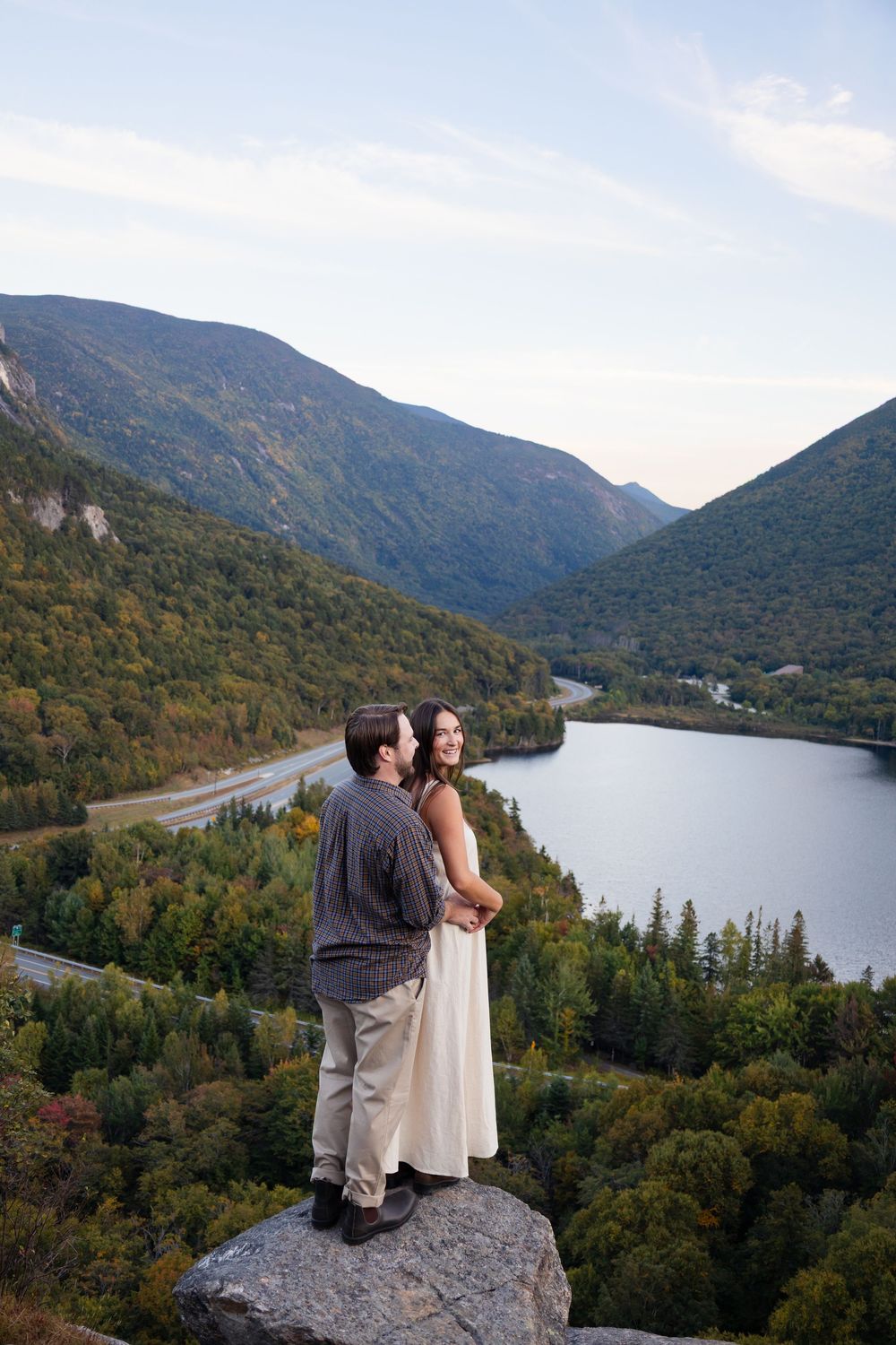 A scenic mountain lake view in autumn with a couple standing on a rocky overlook at Artists Bluff in Franconia Notch.