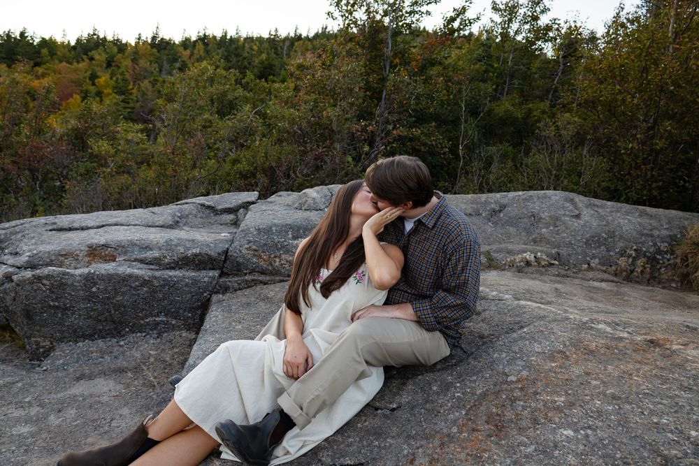 A couple sits intimately on rocky terrain overlooking a scenic forest landscape during an engagement photoshoot.