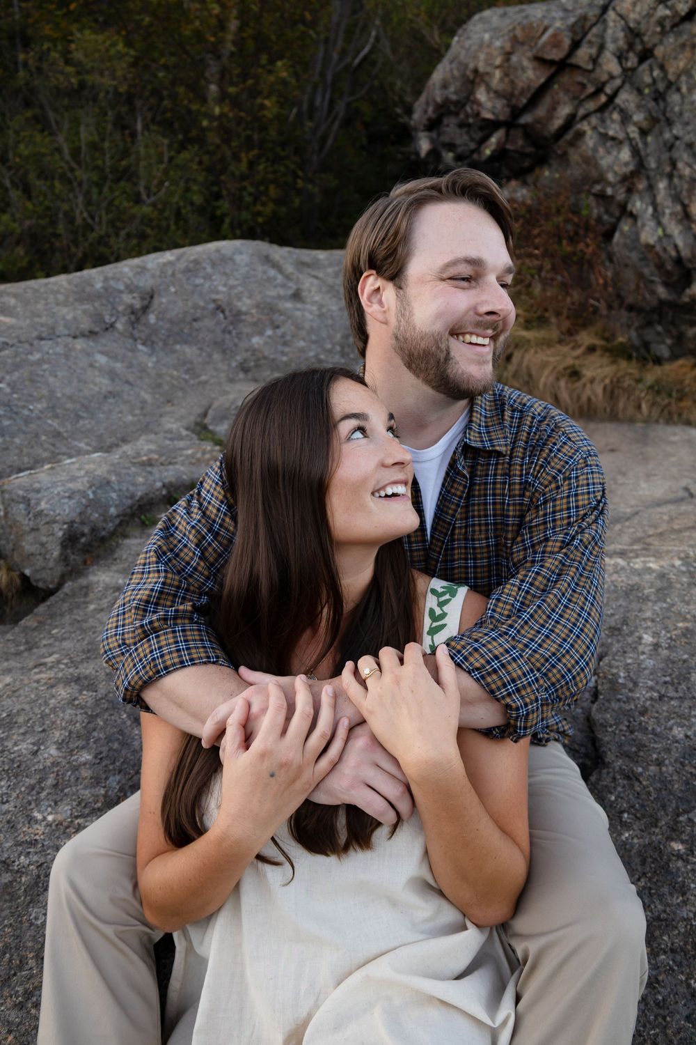 A couple shares joyful laughter while sitting together on rocky terrain during an outdoor photoshoot.