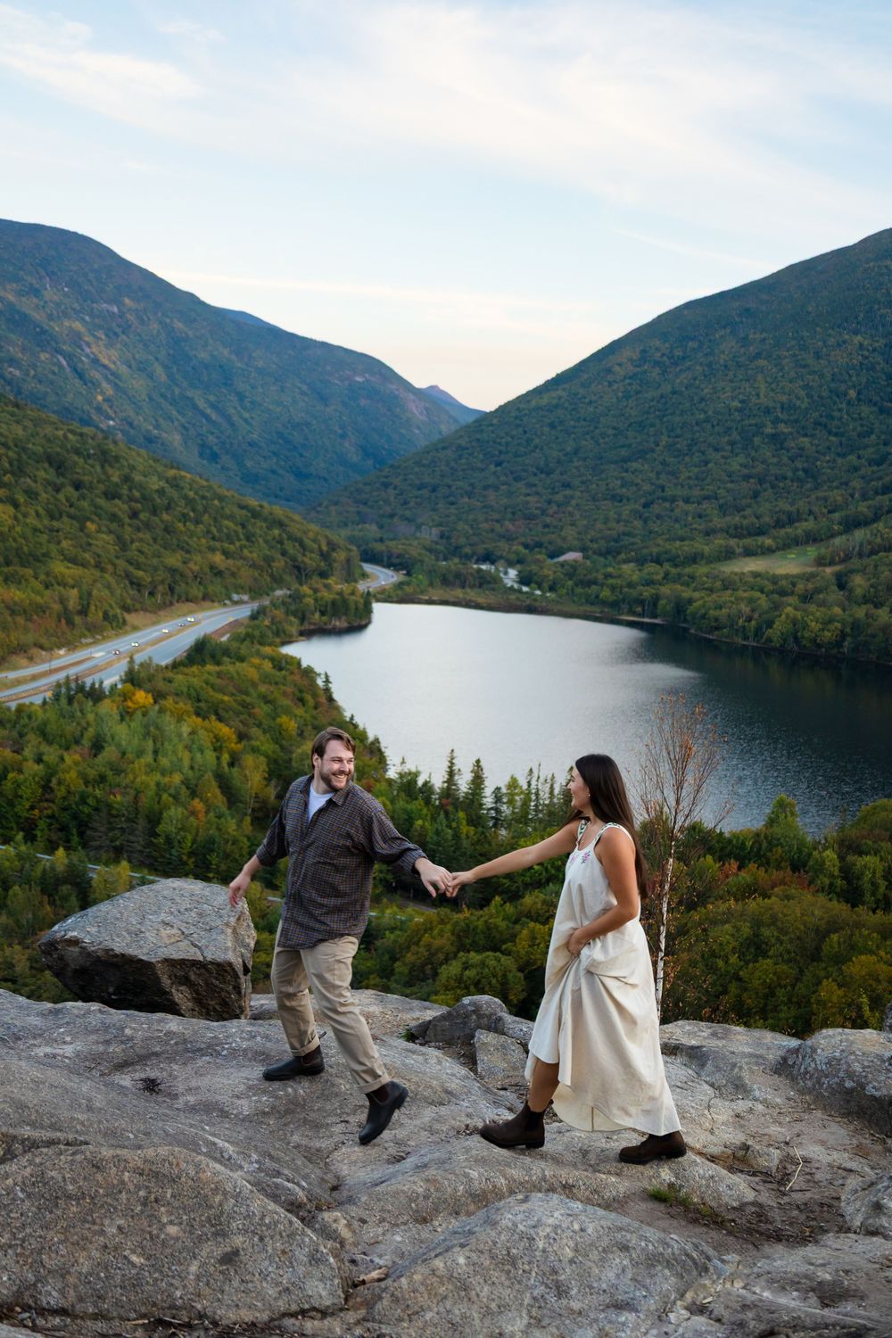 Two people playfully pose on rocky mountain overlook with scenic lake and mountain views in autumn.