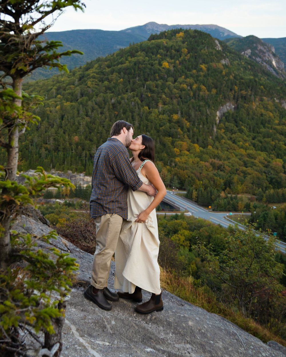 A couple shares a romantic moment on a rocky overlook with mountain vistas and autumn foliage in the background.