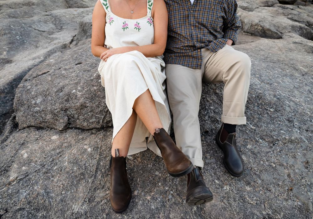 Two people sit together on a rocky surface wearing contrasting white and brown casual outfits and boots.