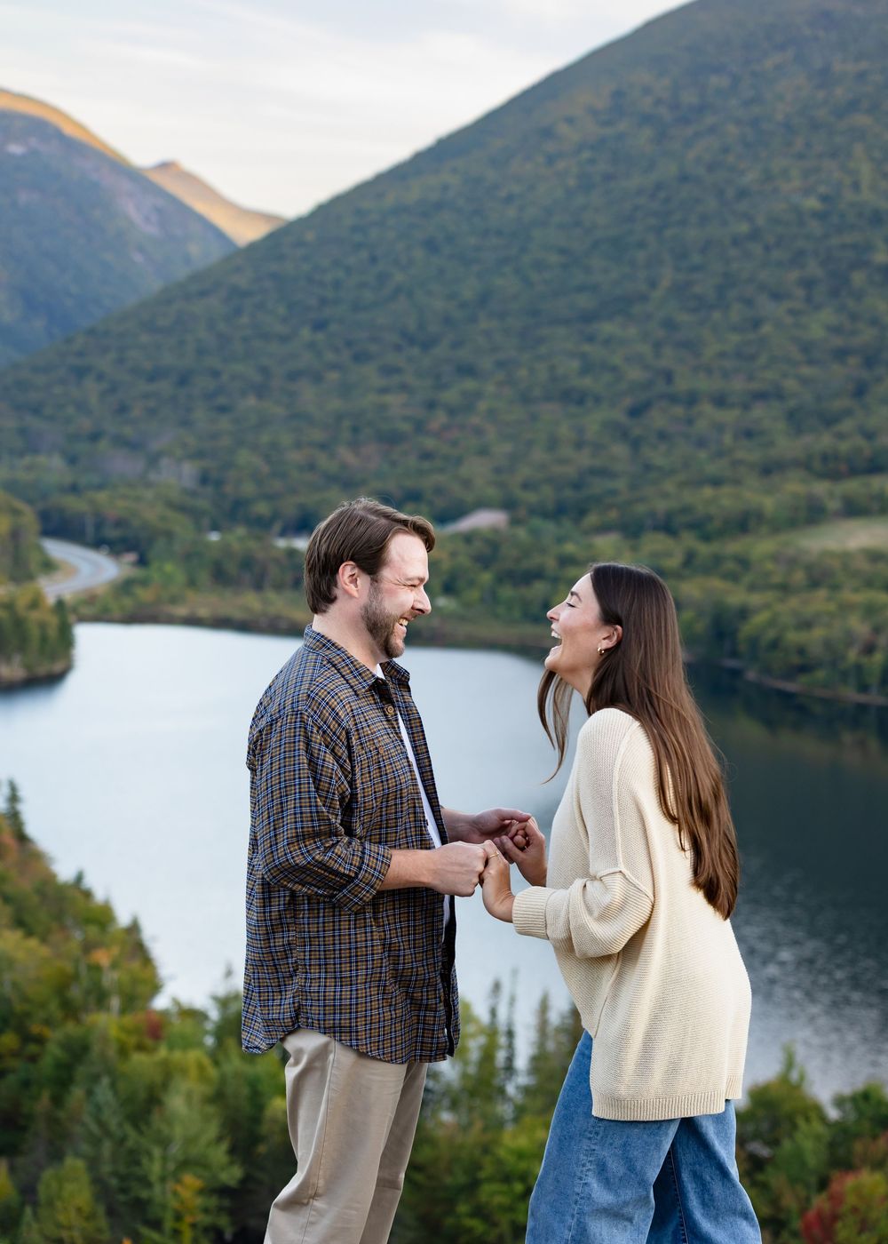 A couple shares a joyful moment while laughing together against a scenic mountain lake backdrop during autumn.