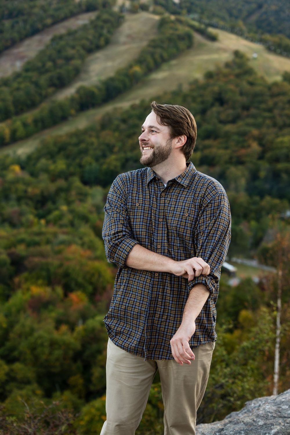 Person in plaid shirt stands on mountain overlook with autumn foliage and ski slopes visible in background.