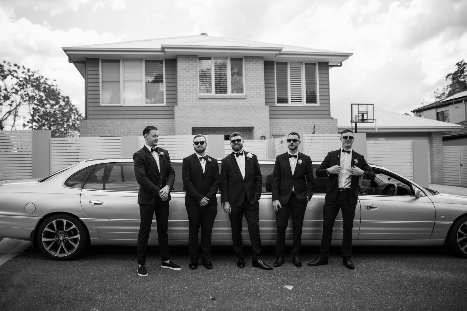 A black and white panoramic photo showing a group of people in formal attire standing beside a luxury vehicle.