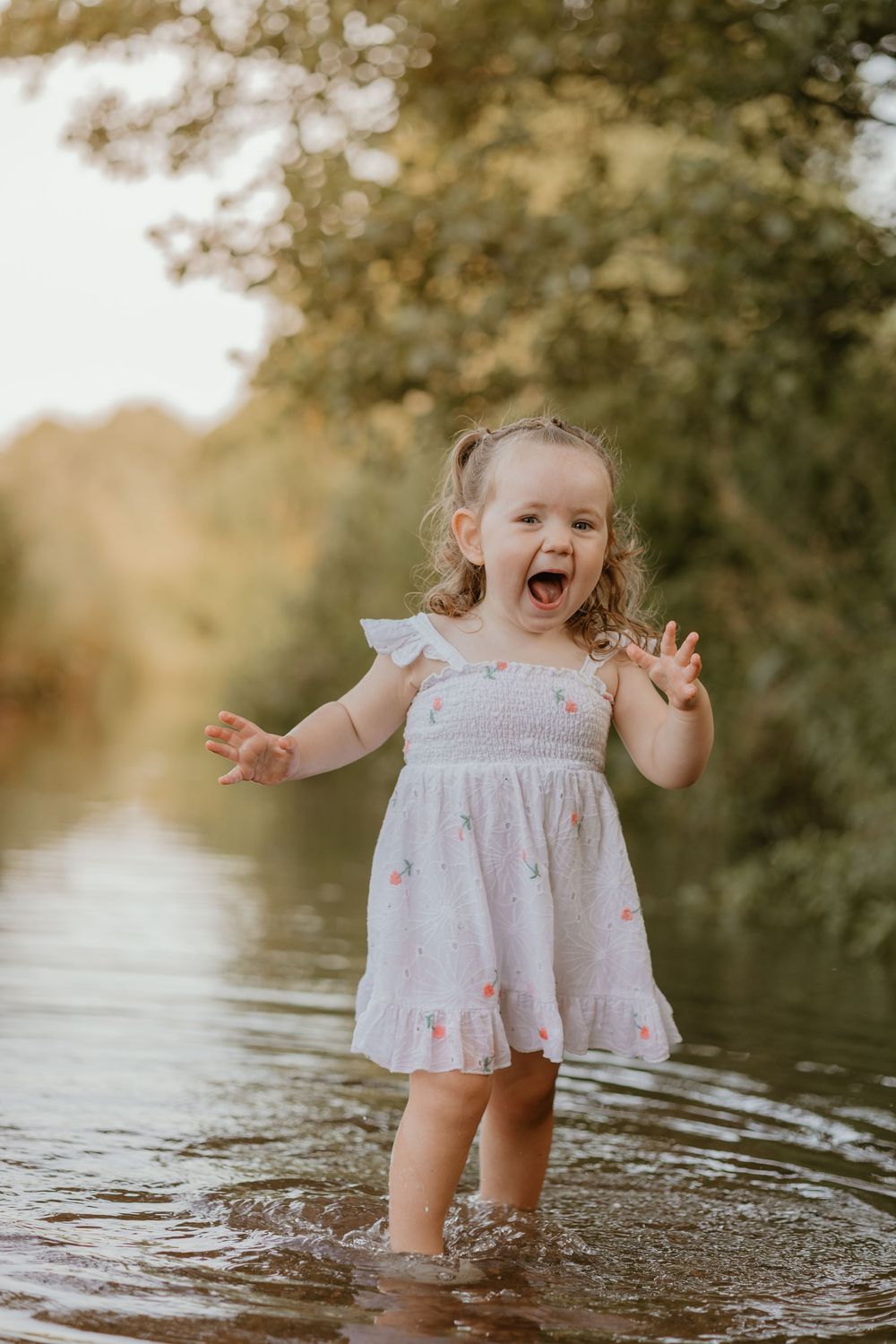 A small girl in a white dress splashes and plays in shallow water during sunset.
