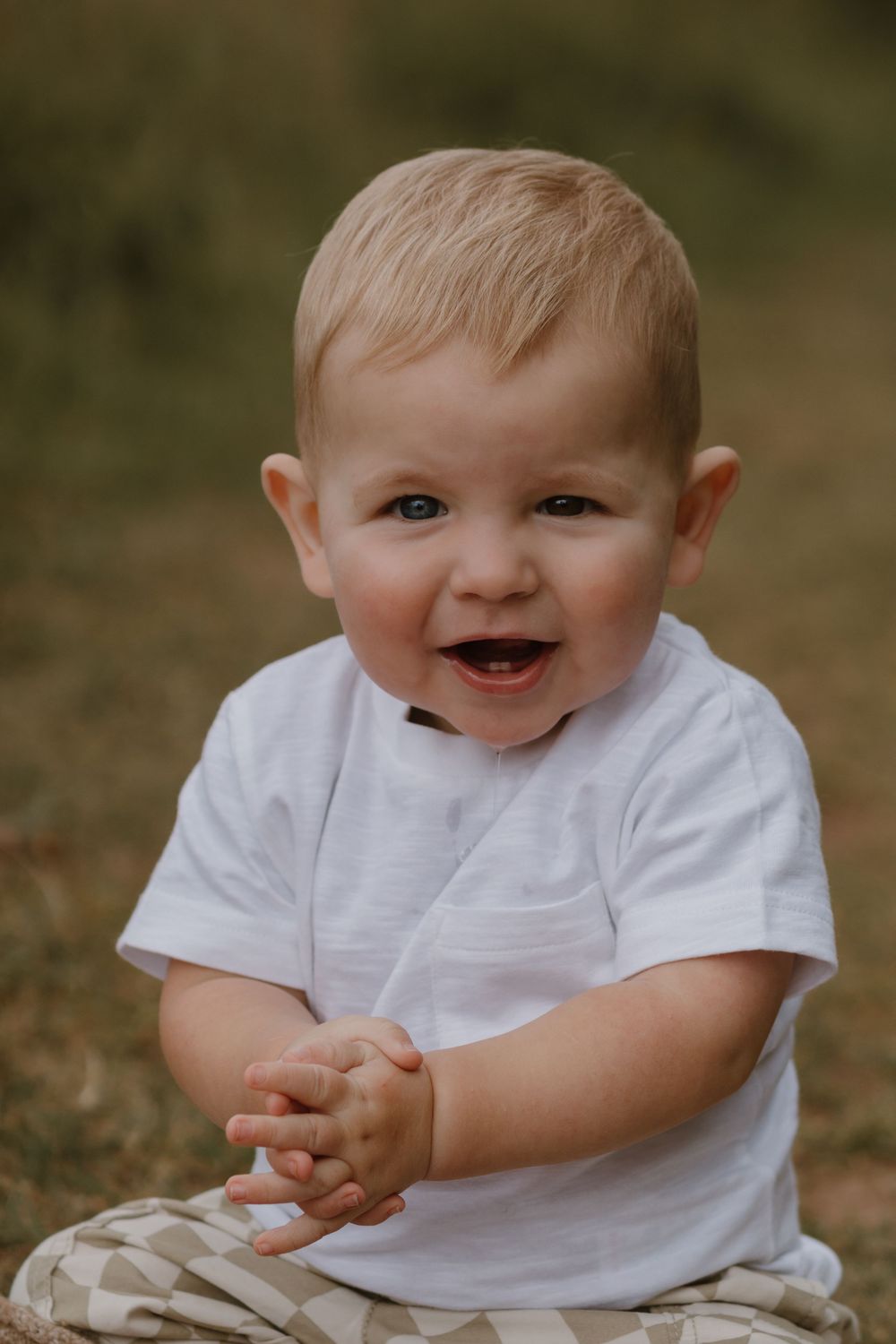 A baby in white clothing smiles and plays while sitting outdoors.