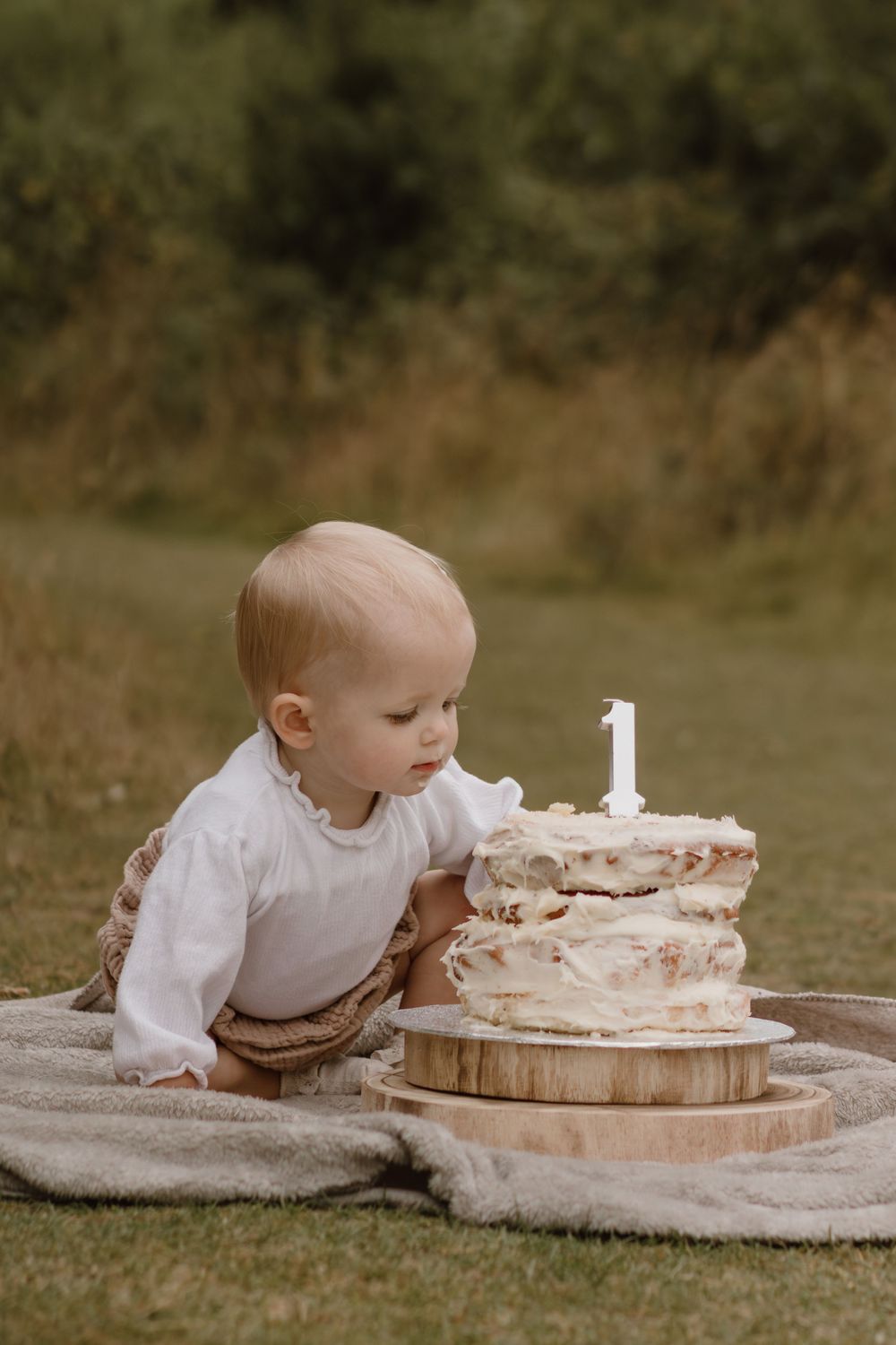 A baby leans towards a white birthday cake with number one candle on a blanket outdoors.