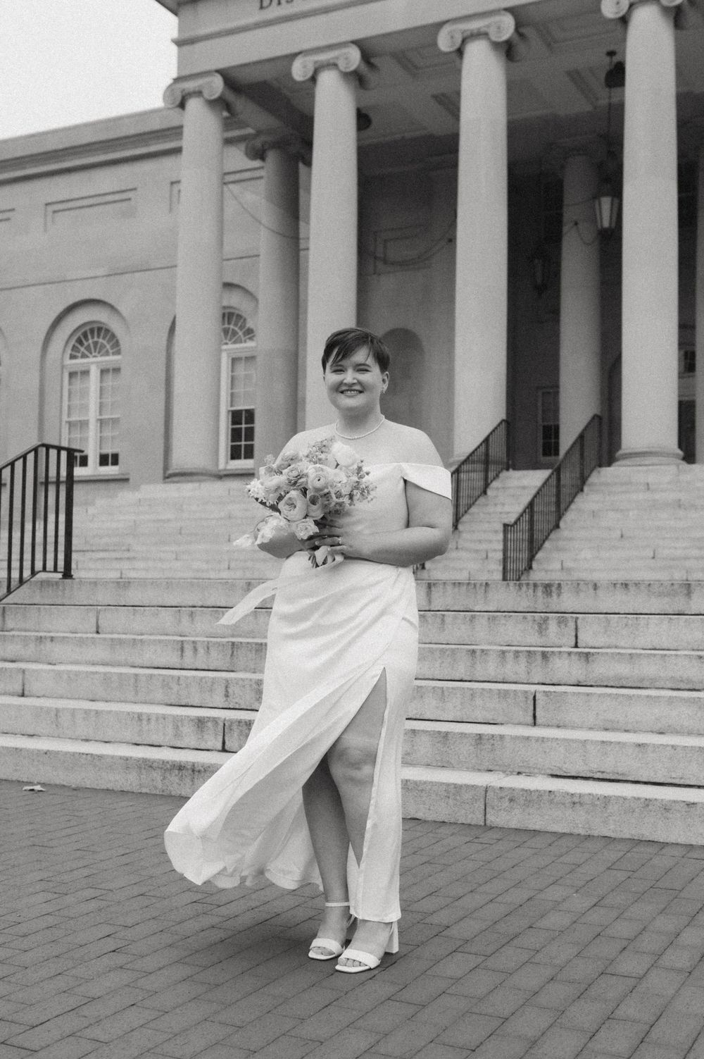 Figure in white outfit poses playfully on steps of a classical building with columns.