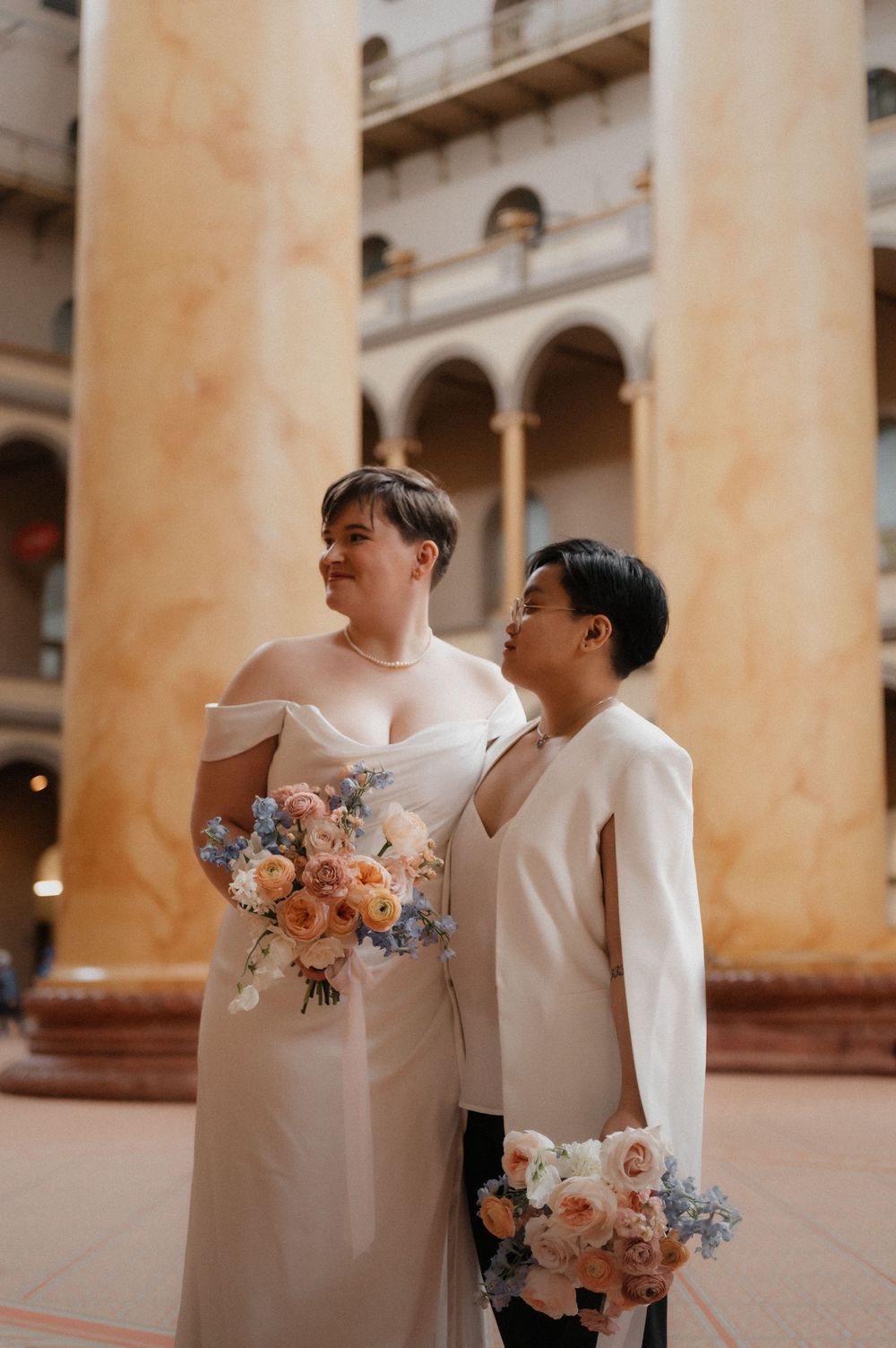 Couple posing between elegant stone columns holding peach and blue floral bouquets.