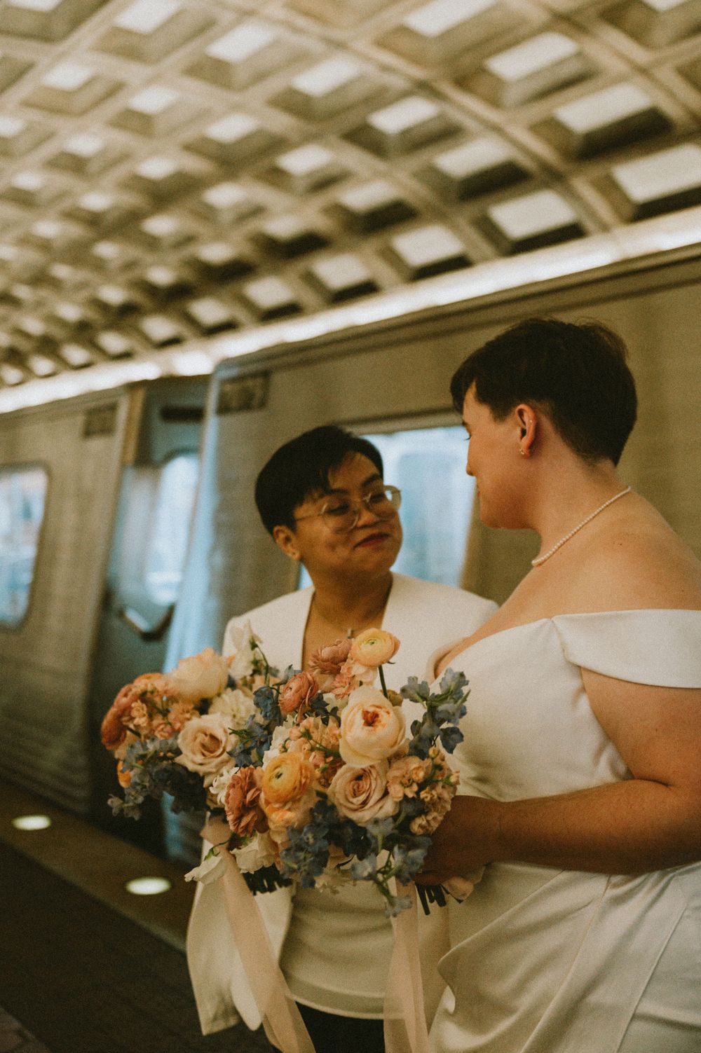 Wedding party members in white attire hold peach and blue floral bouquets in a metro station.
