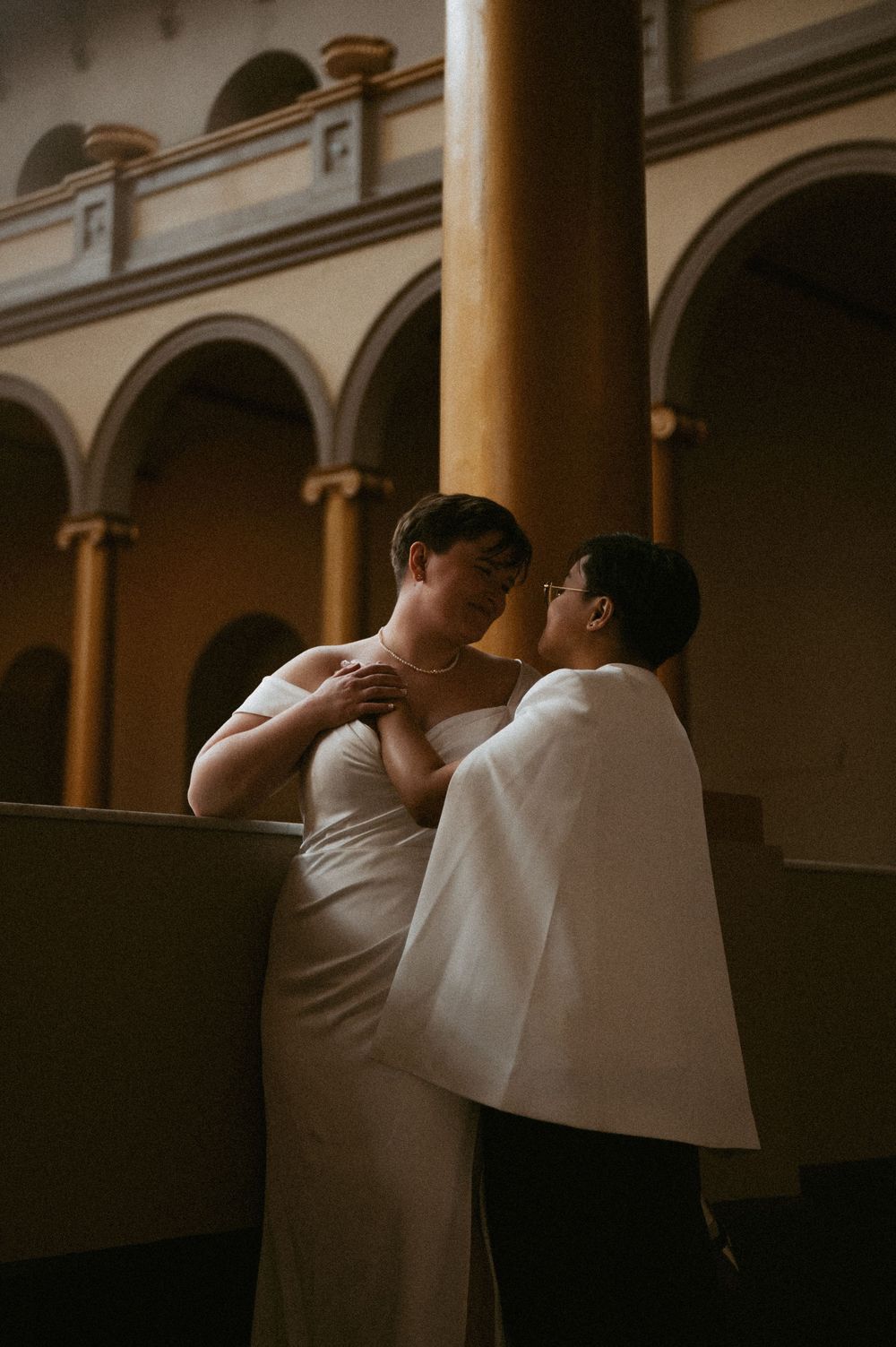 A romantic couple shares an intimate moment in an elegant arched corridor with classical architecture.