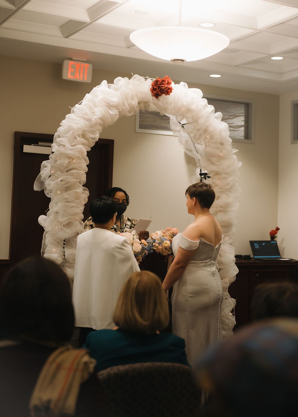 Wedding ceremony sequence under a white floral arch in an indoor venue with guests watching.