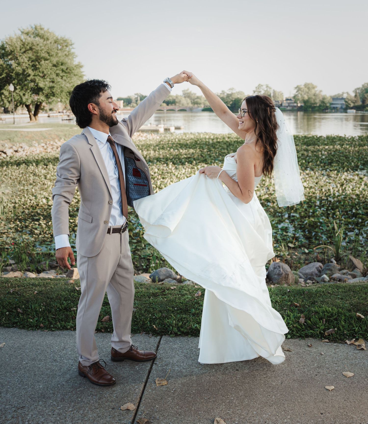 Bride and groom dancing outdoors near a lake, groom in tan suit twirling bride in white gown.