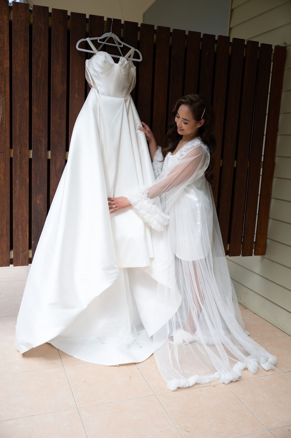 Bride in a white robe admiring her off-shoulder wedding gown hanging on a wooden wall.