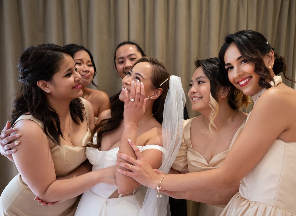 Excited bride surrounded by bridesmaids in champagne dresses sharing a joyful wedding moment.