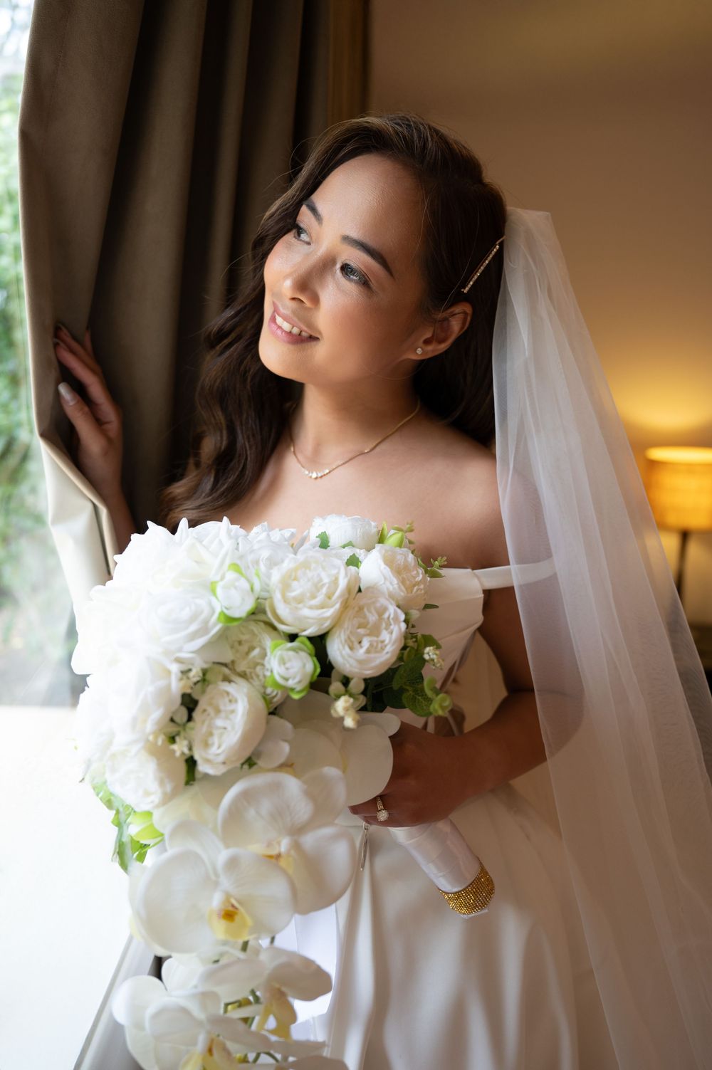 Radiant bride in a white strapless gown holding a lush white bouquet with orchids by a window.