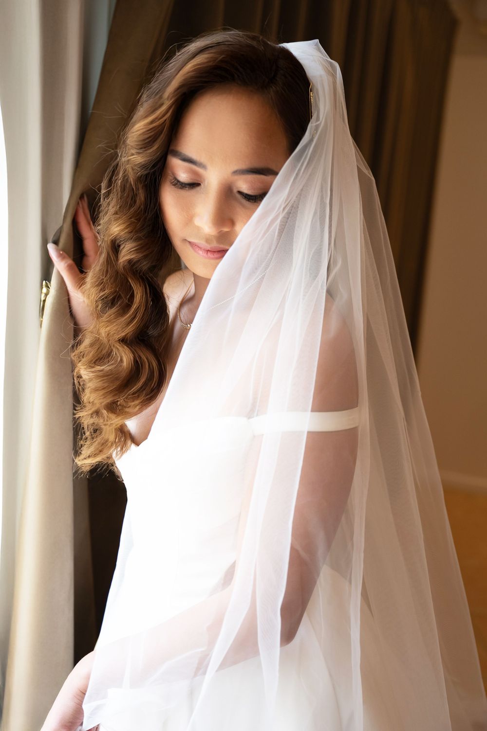 Elegant bride with flowing veil draped over her face posing softly by a window in white gown.