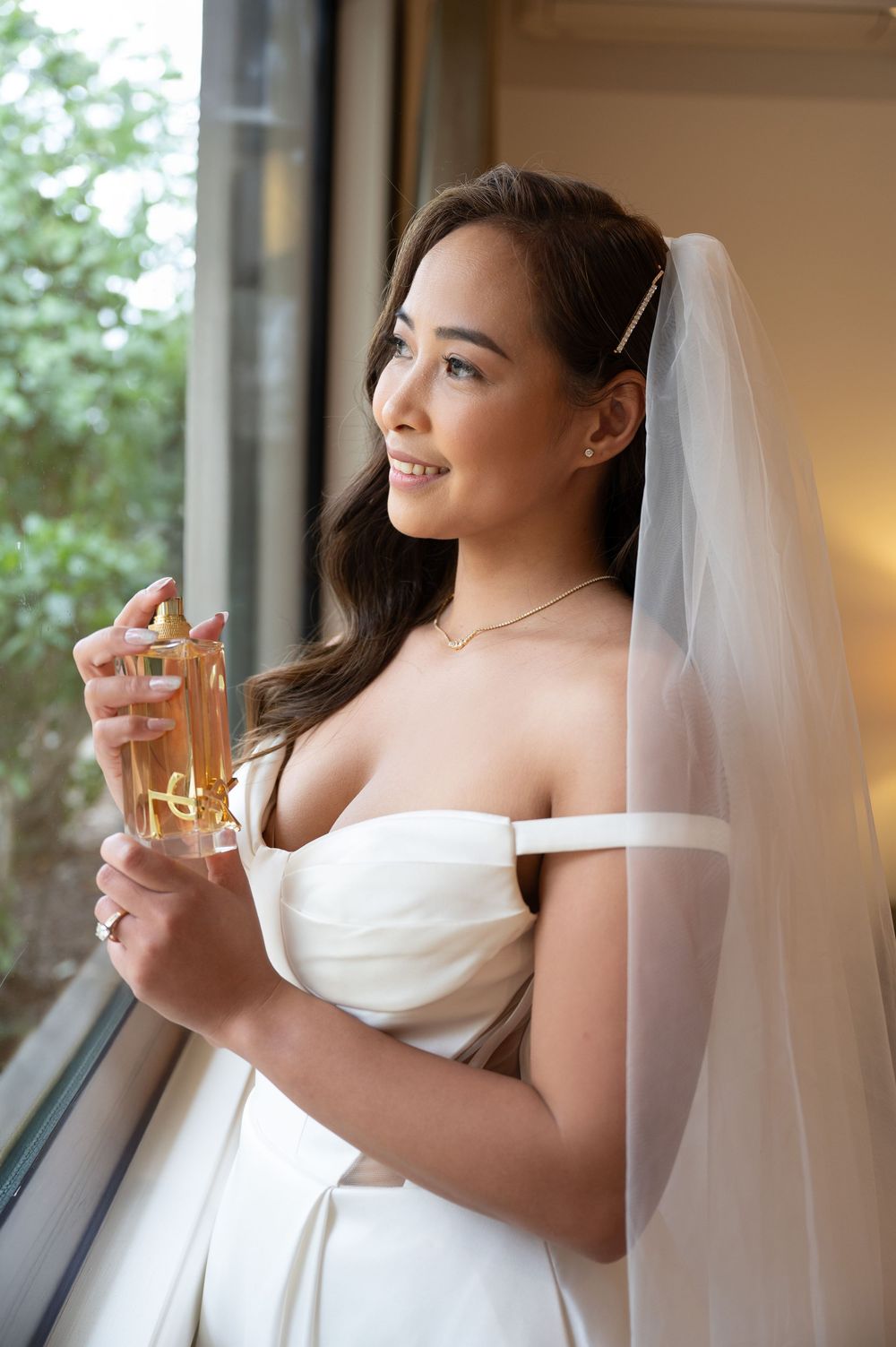 Beautiful bride in off-shoulder wedding dress holding perfume bottle while gazing out a window.