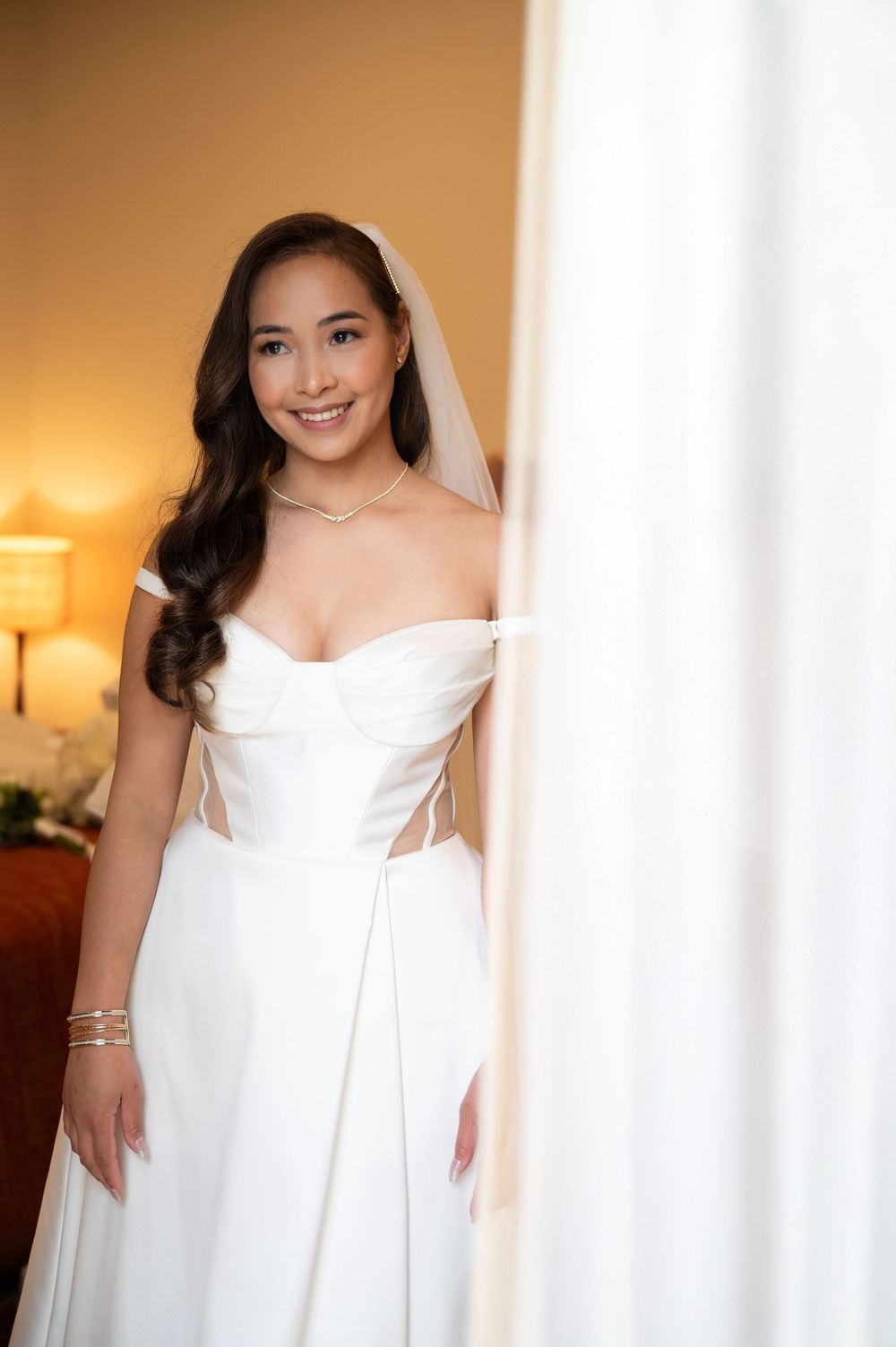 Smiling bride in a structured off-shoulder white wedding gown with veil standing by a curtain.