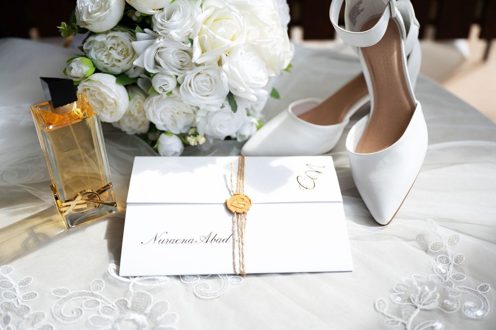 Elegant wedding flat lay with white heels, invitation, perfume bottle, and white rose bouquet.