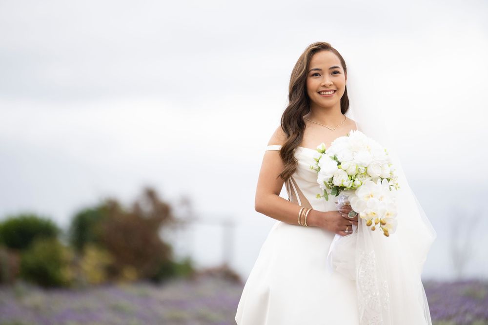 Smiling bride in white off-shoulder gown holding white bouquet in lavender field.