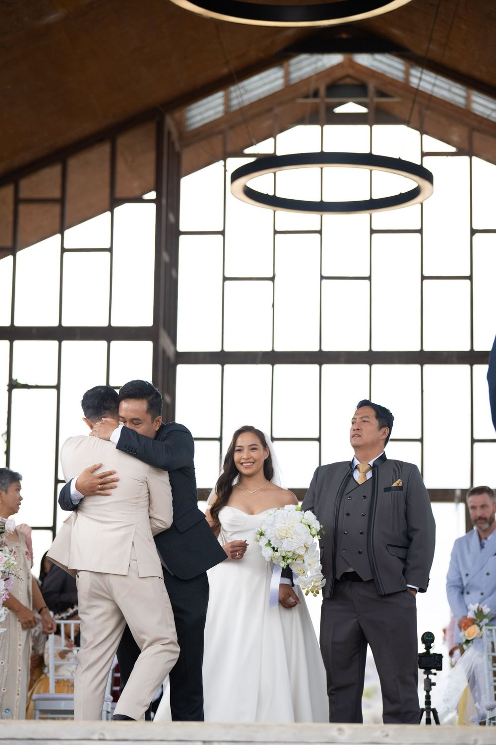 Groom hugging guest while bride smiles during wedding ceremony in rustic venue.