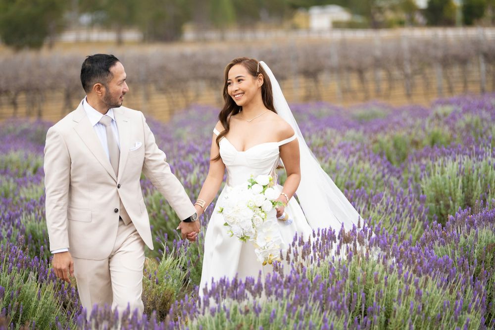 Bride and groom walk hand in hand smiling through blooming purple lavender field with white bouquet.
