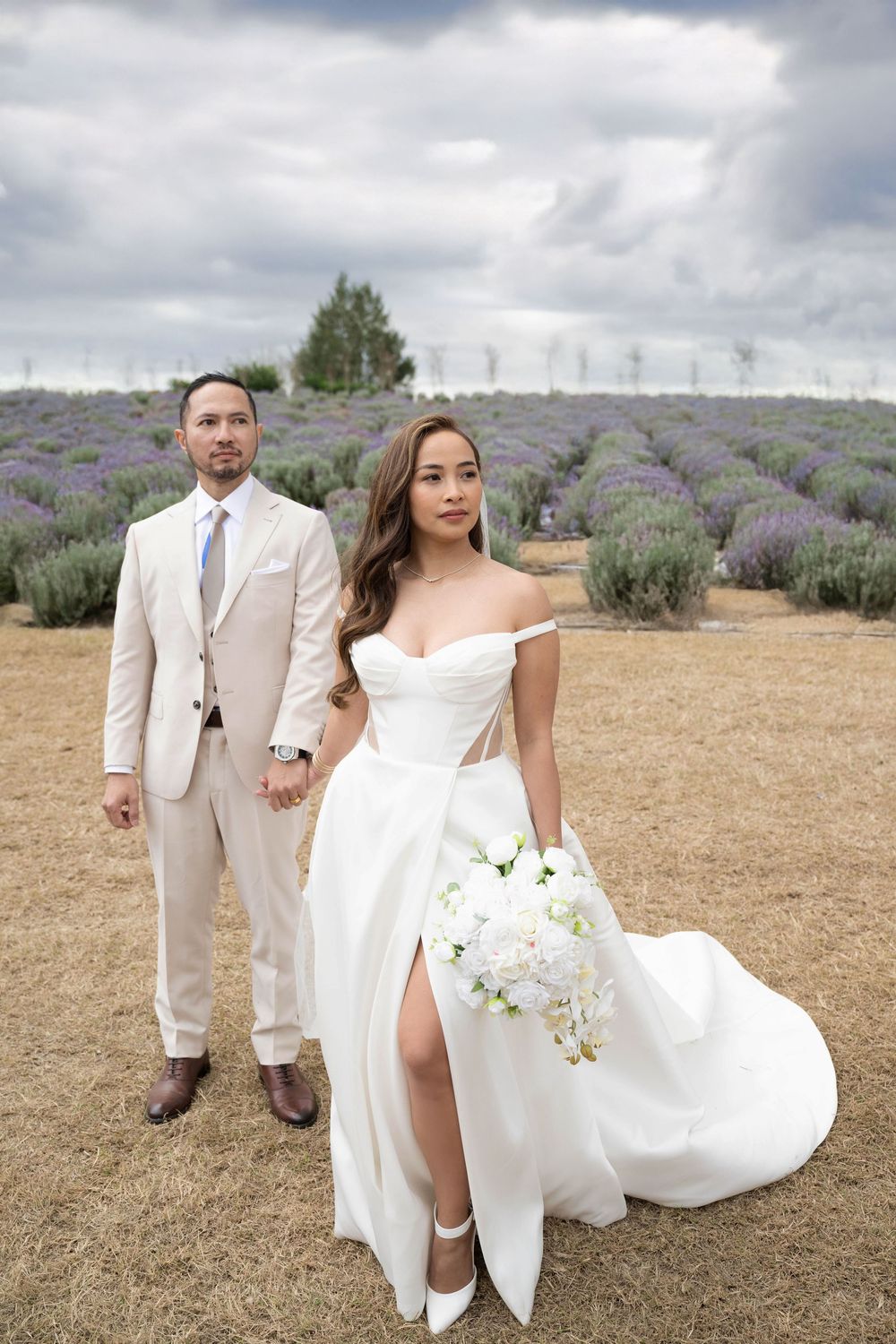 Elegant bride in off-shoulder gown holds bouquet while groom stands beside her in lavender field.