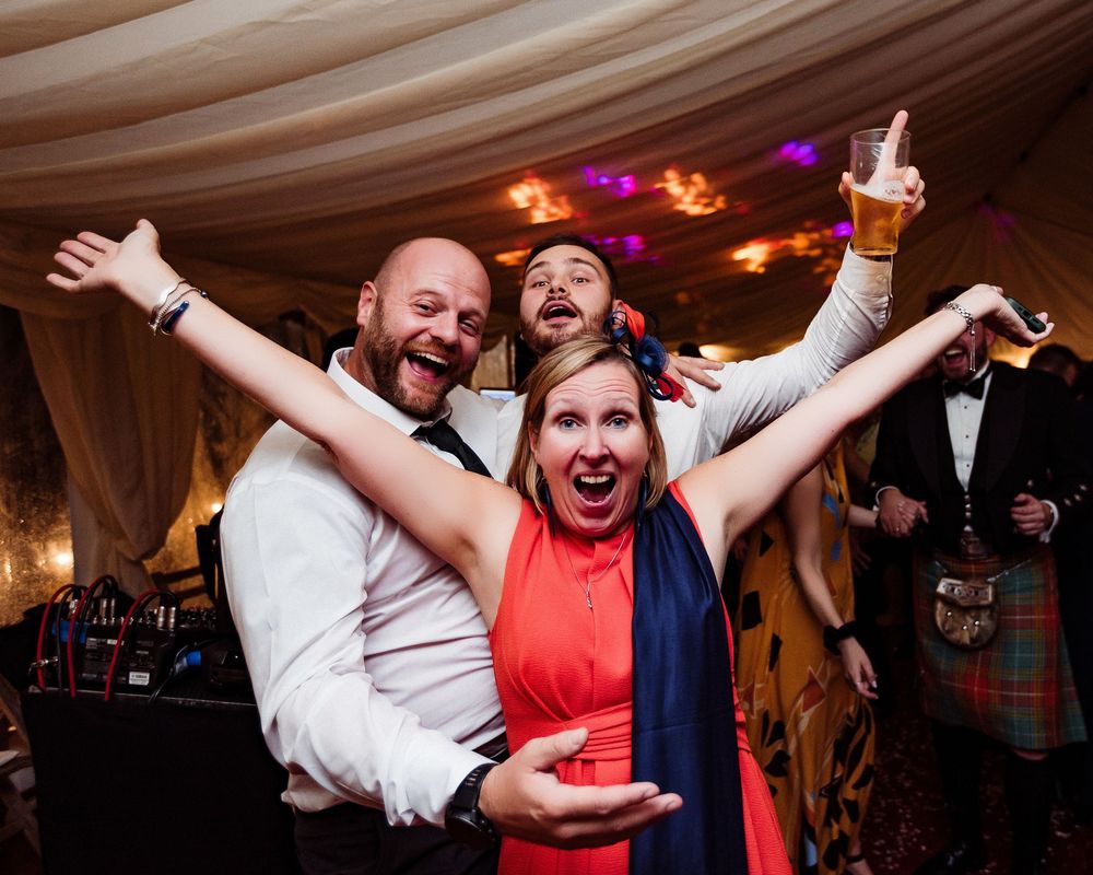 People dancing and celebrating with energetic movements at a wedding reception on a dark dance floor with colored lights.