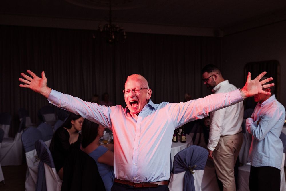 People dancing and celebrating with raised arms on a dimly lit dance floor at a wedding reception.