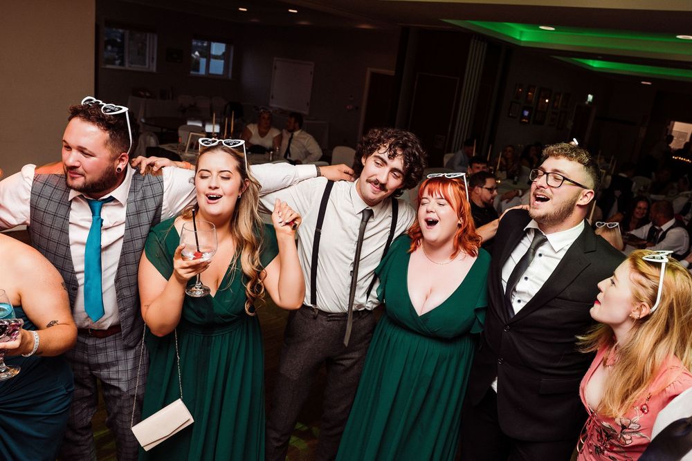 Wedding party guests in emerald green dresses and formal attire laugh and dance together at a reception celebration.