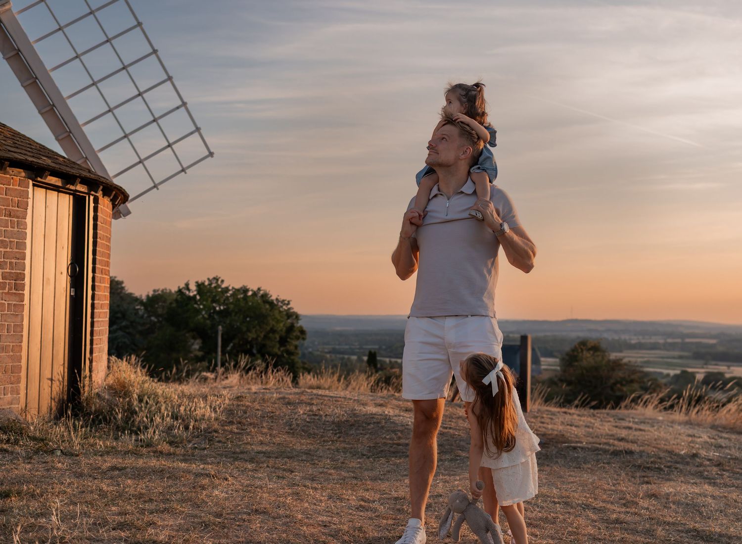Father carries daughter on shoulders near a windmill at sunset, while another child stands beside them.