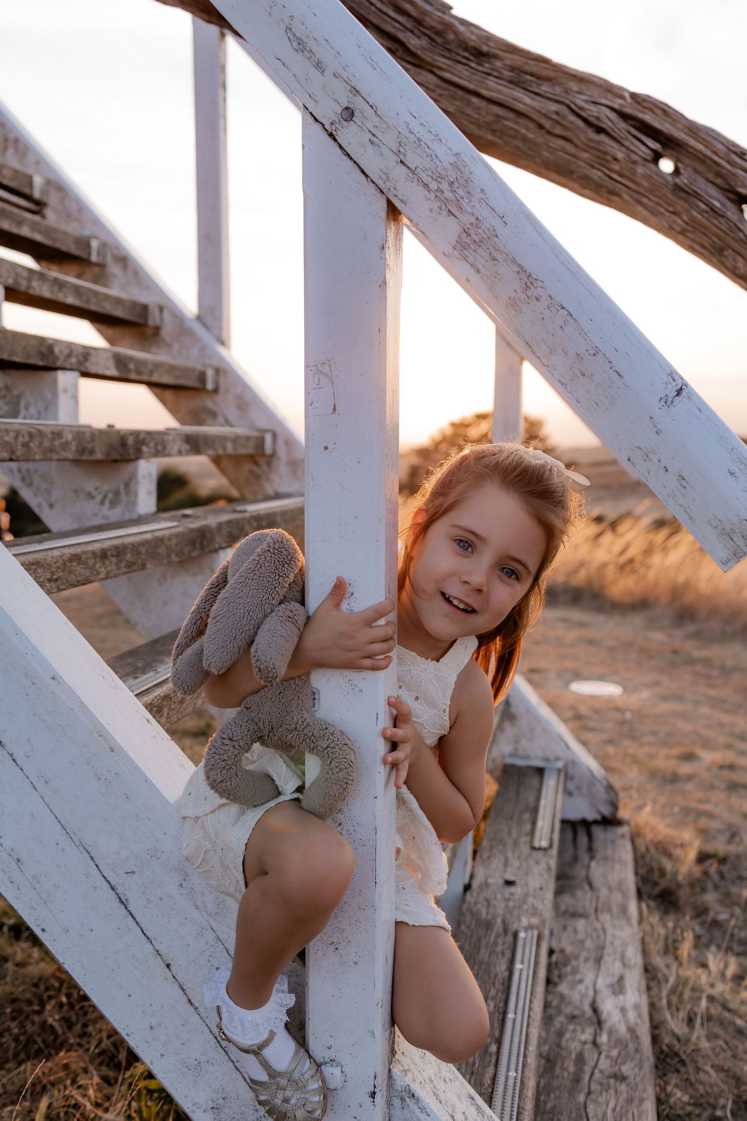 Young girl in white dress hugging stuffed bunny on rustic white wooden stairs at golden hour sunset.