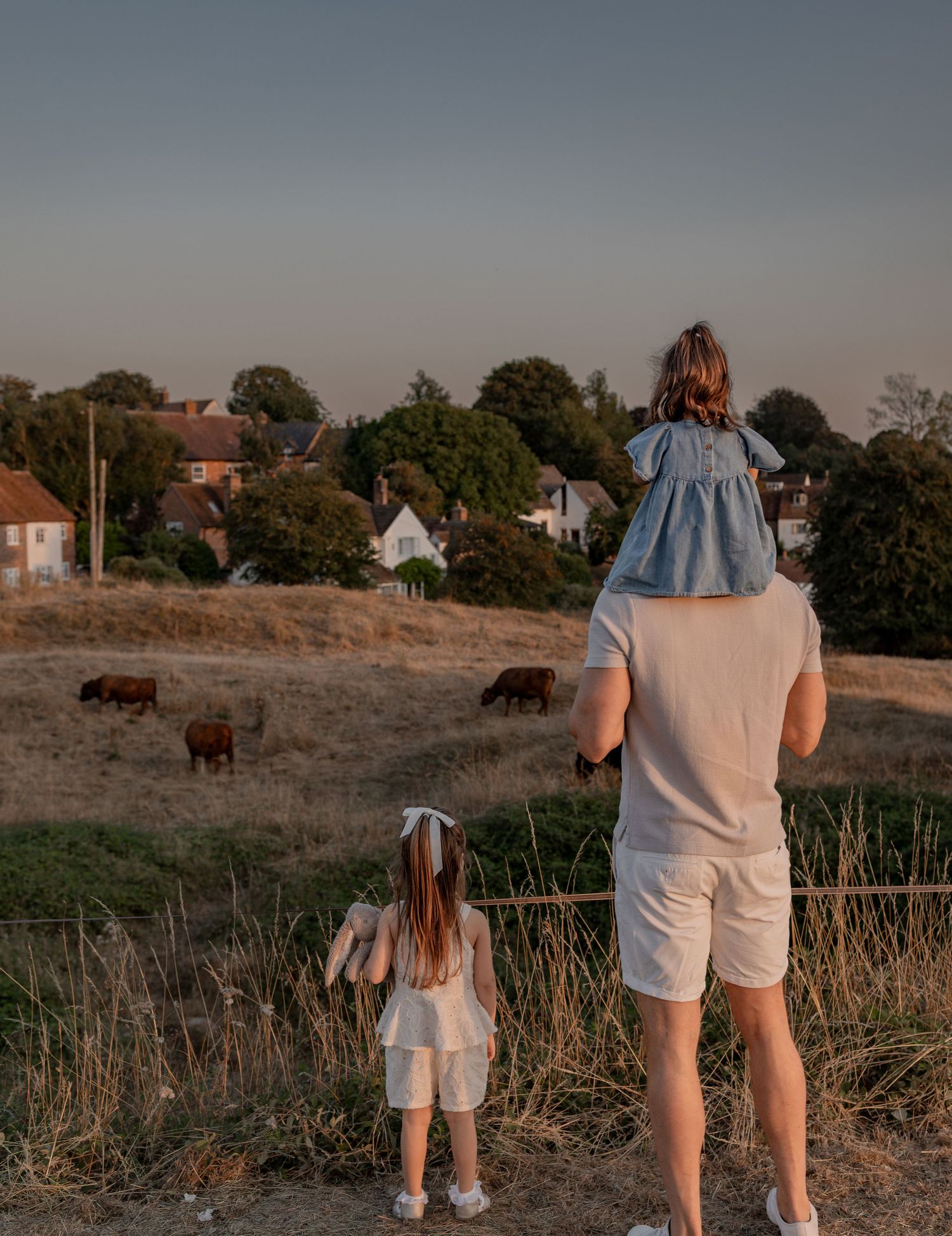 A father holds his daughter on his shoulders while another child stands beside him, overlooking grazing cows and a village.